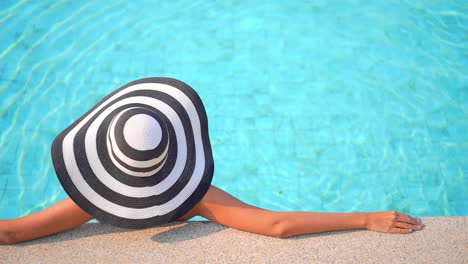 back of a woman in floppy stripped hat leaning on pool border inside swimming pool, template copy space