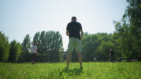 Back view of a man rotating a soccer ball around him while others play in the background, during a sunny day with trees and a goalpost in the distance