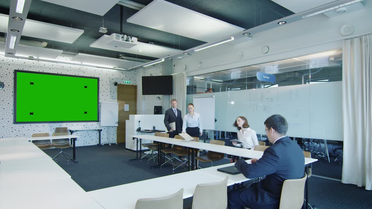 사무실 직원들이 회의실로 오고 있습니다. 벽에는 녹색 화면이 표시되어 있습니다.