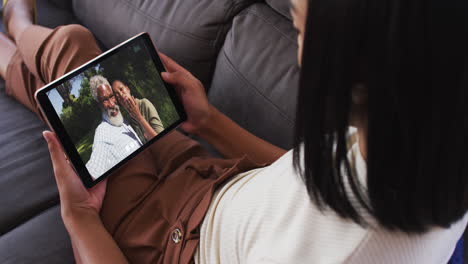 mujer biracial sentada en el sofá usando una tableta haciendo una llamada de video con la familia
