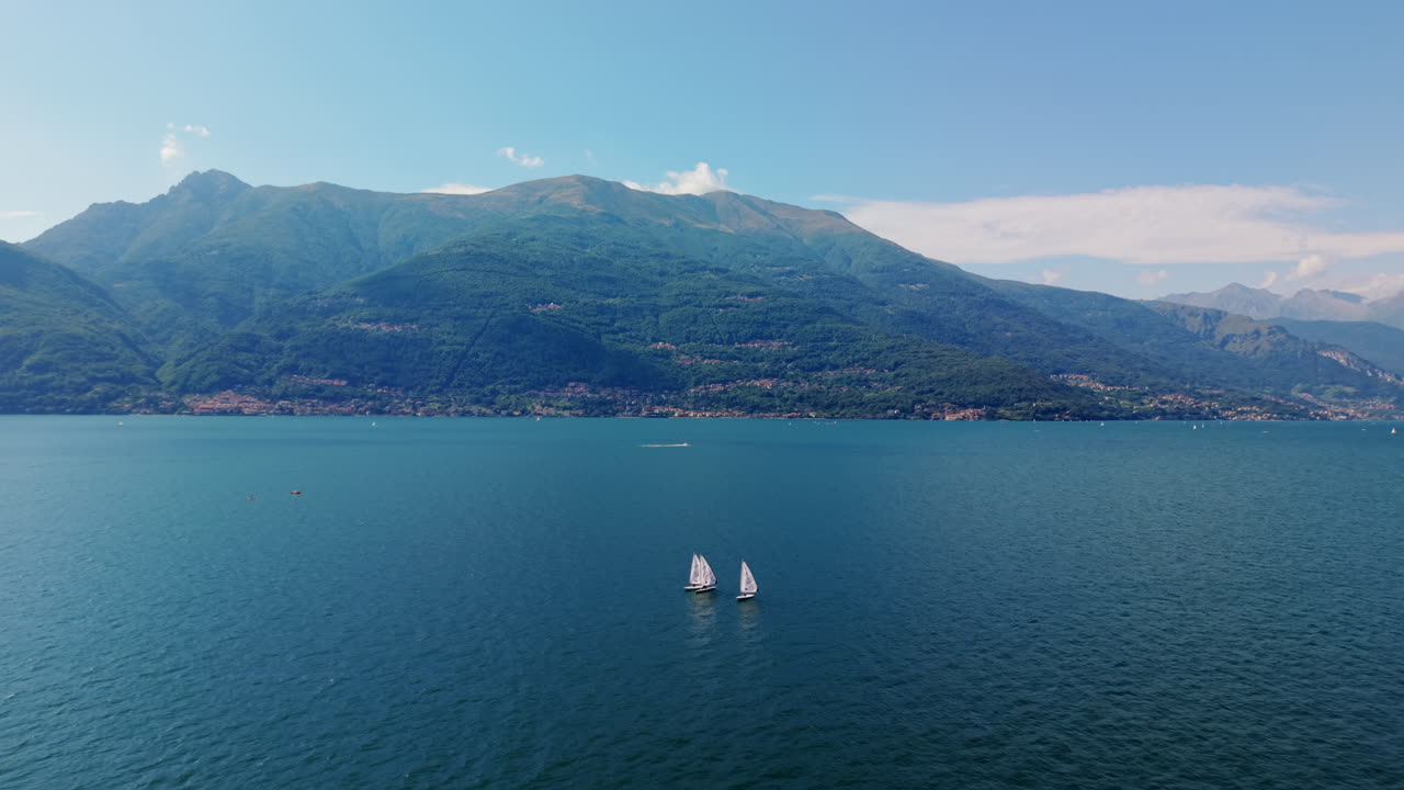 Drone flies forward at steady height toward three small sailboats on a wide lake, with distant mountain slopes visible in the background