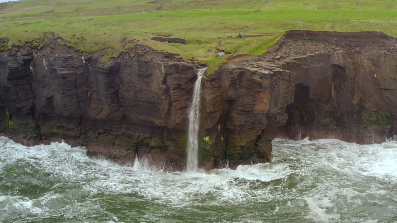 Aerial orbit of waterfall dropping into roaring Atlantic at Cliffs of Moher in Doolin, with hikers passing and a stone bridge slowly revealed