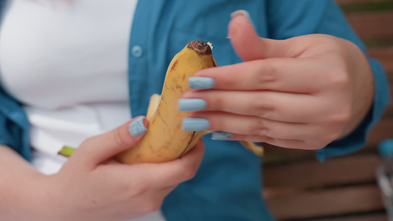 primer plano de una joven pelando la piel de un plátano con ambas manos, la luz suave del día resaltando la textura natural de la fruta y el esmalte de uñas azul, sentada al aire libre en un banco de madera