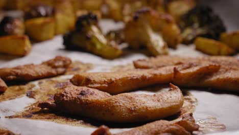 a close up shot tilting down to reveal marinated chicken strips on a tray covered in parchment paper, the tray shared with an assortment of vegetables as they roast in a hot kitchen oven