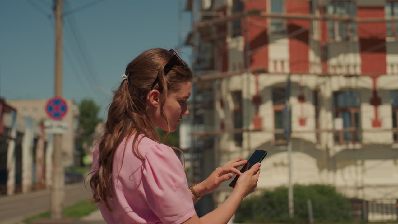 back view caucasian woman walking street, checking phone with red scaffolding backdrop, casual pink dress, long braid, tourist curiosity, warm light, slow pace, downtown renovation context