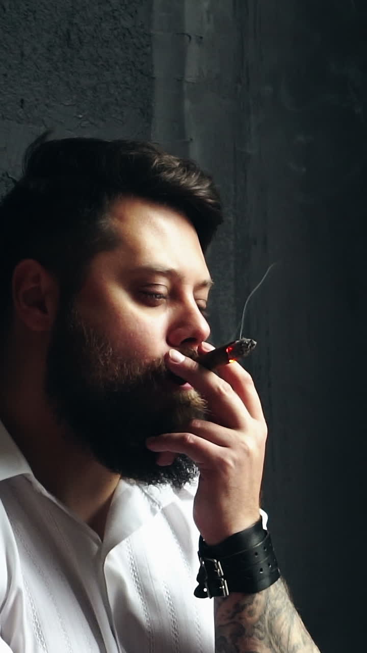Brutal handsome man with cigar. Young brutal man relaxing and smoking cigar in studio Vertical video