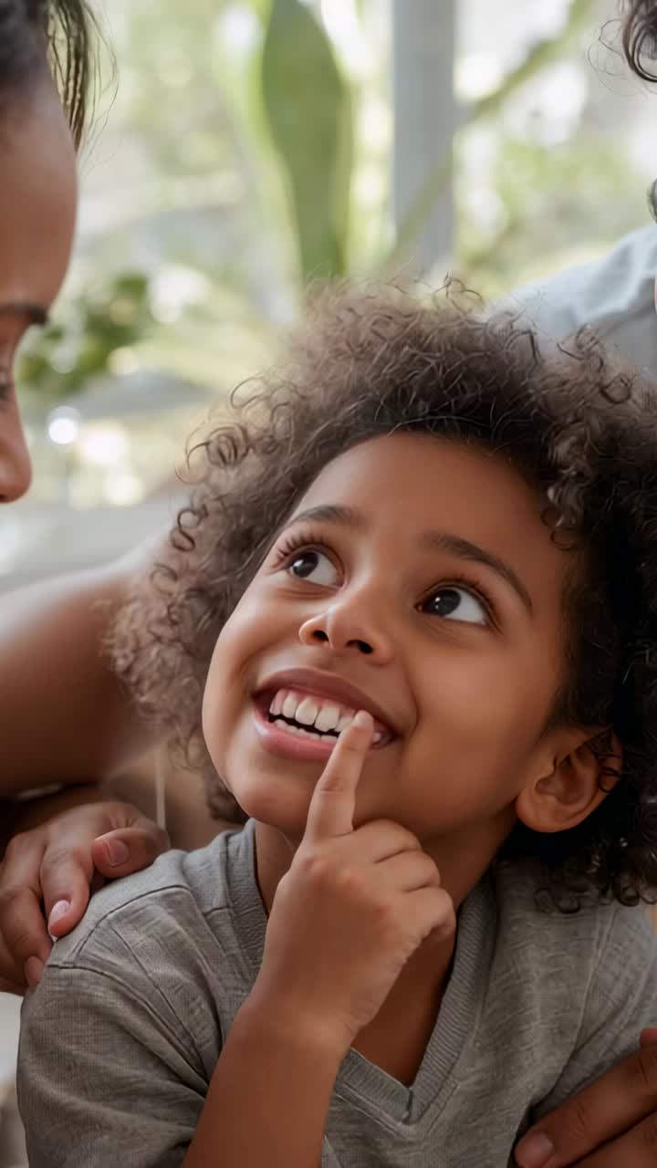 Vertical video: Smiling child in gray tee touching tooth on couch by window, adults leaning in