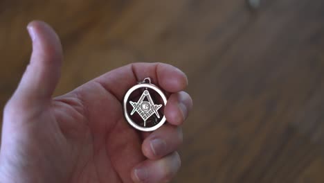 Caucasian-white-man’s-hands-holding-Masonic-G-symbol-talisman-necklace-in-hand,-with-wooden-chopping-block-table-in-background