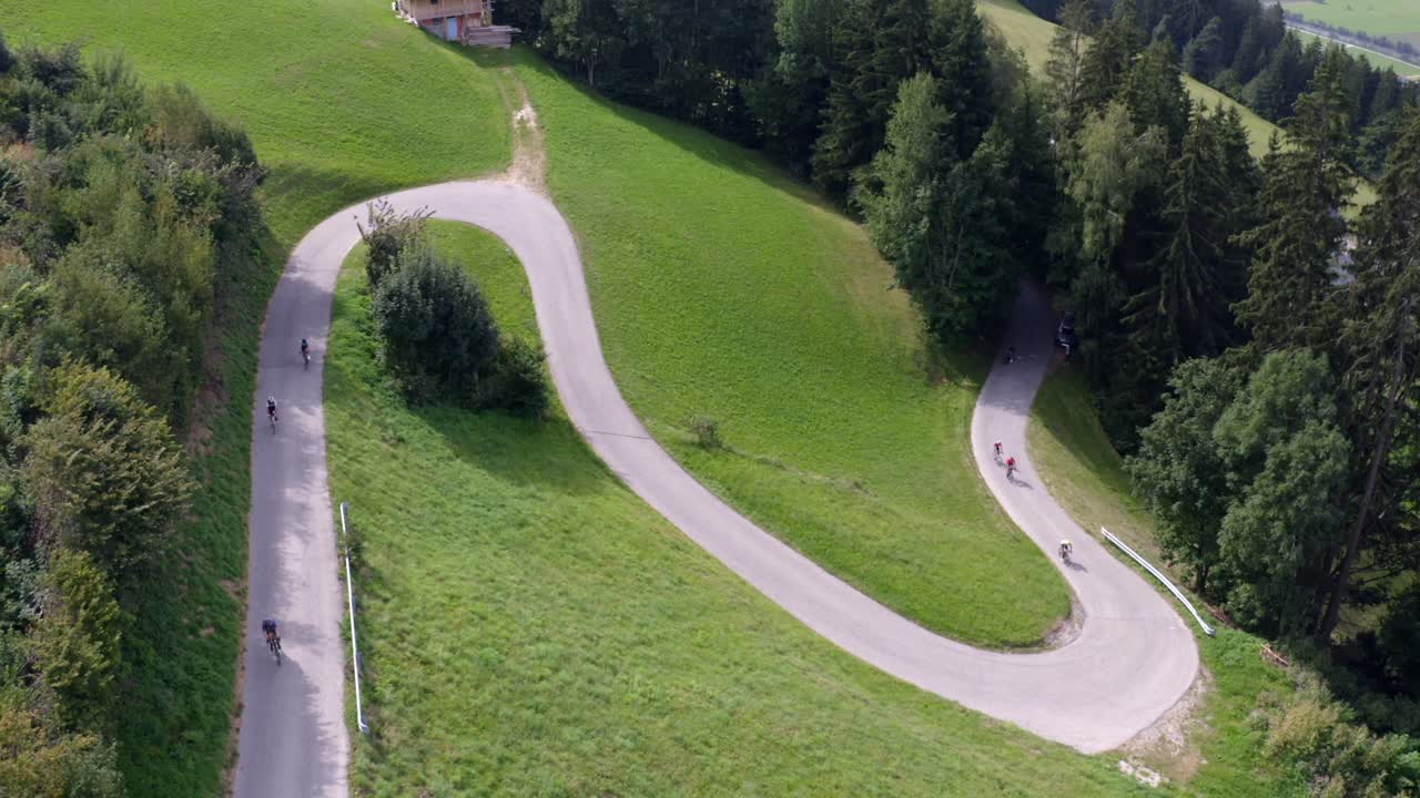 Cyclists making their way through a serpentine country road amid green hills and scattered trees.