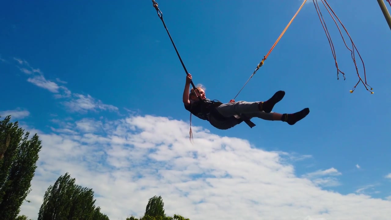 Un'inquadratura statica dal basso cattura un bambino imbracato in un sistema elastico di bungee trampoline, che salta in alto contro un cielo azzurro brillante con nuvole sparse a un festival del parco