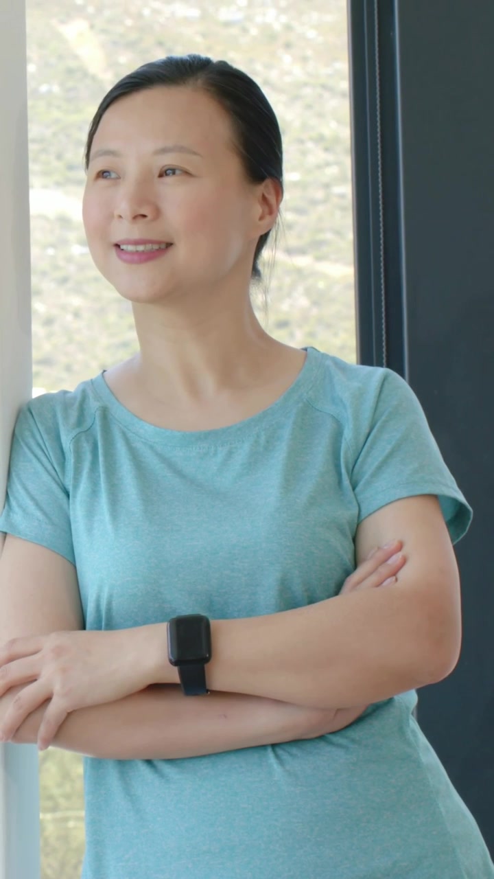 縦型動画：窓際でティール色のTシャツと黒い時計を身につけた女性が、光に誘われて丘陵地帯に視線を向けている様子。