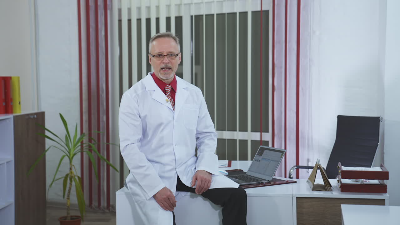 Medical specialist giving online consultation. Friendly senior doctor in white coat and glasses sitting on desk in medical room and explaining something on camera.