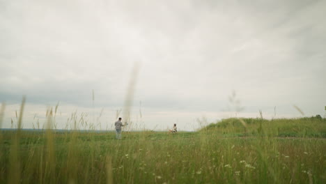 a painter, wearing a hat and checkered shirt, is engaged in painting on an easel in a meadow filled with tall grass, under a cloudy sky. a woman in a hat and white gown sits peacefully in a chair