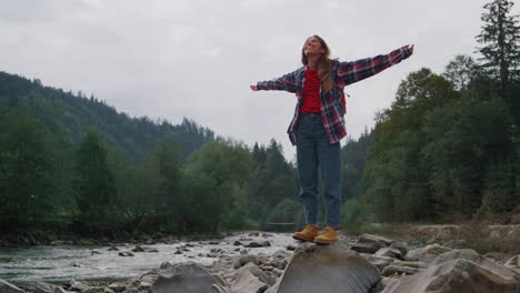 happy female hiker standing at rocky river shore. smiling woman raising hands