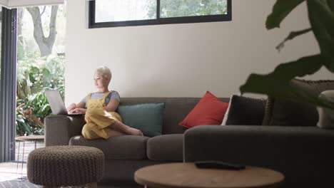 Happy-biracial-woman-using-laptop-and-sitting-on-couch-in-slow-motion