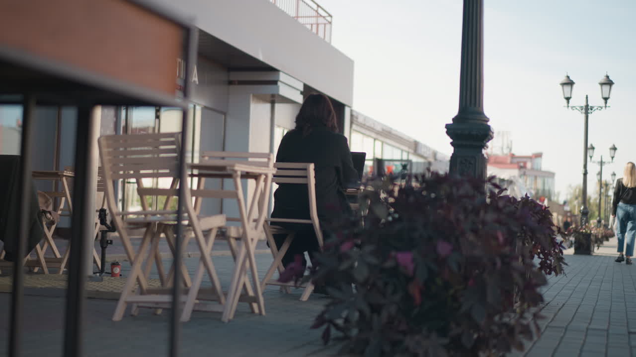 vista trasera de una mujer escribiendo en una computadora portátil, rodeada de sillas de madera y plantas en una cafetería al aire libre, la escena es pacífica con un vistazo de una persona en la distancia caminando