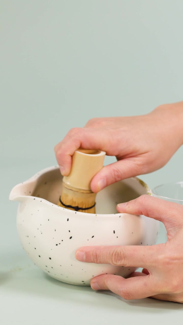 Mani sbattono il tè matcha usando un frullino di bambù in una ciotola di ceramica, illuminazione soffusa, sfondo pulito, primo piano.