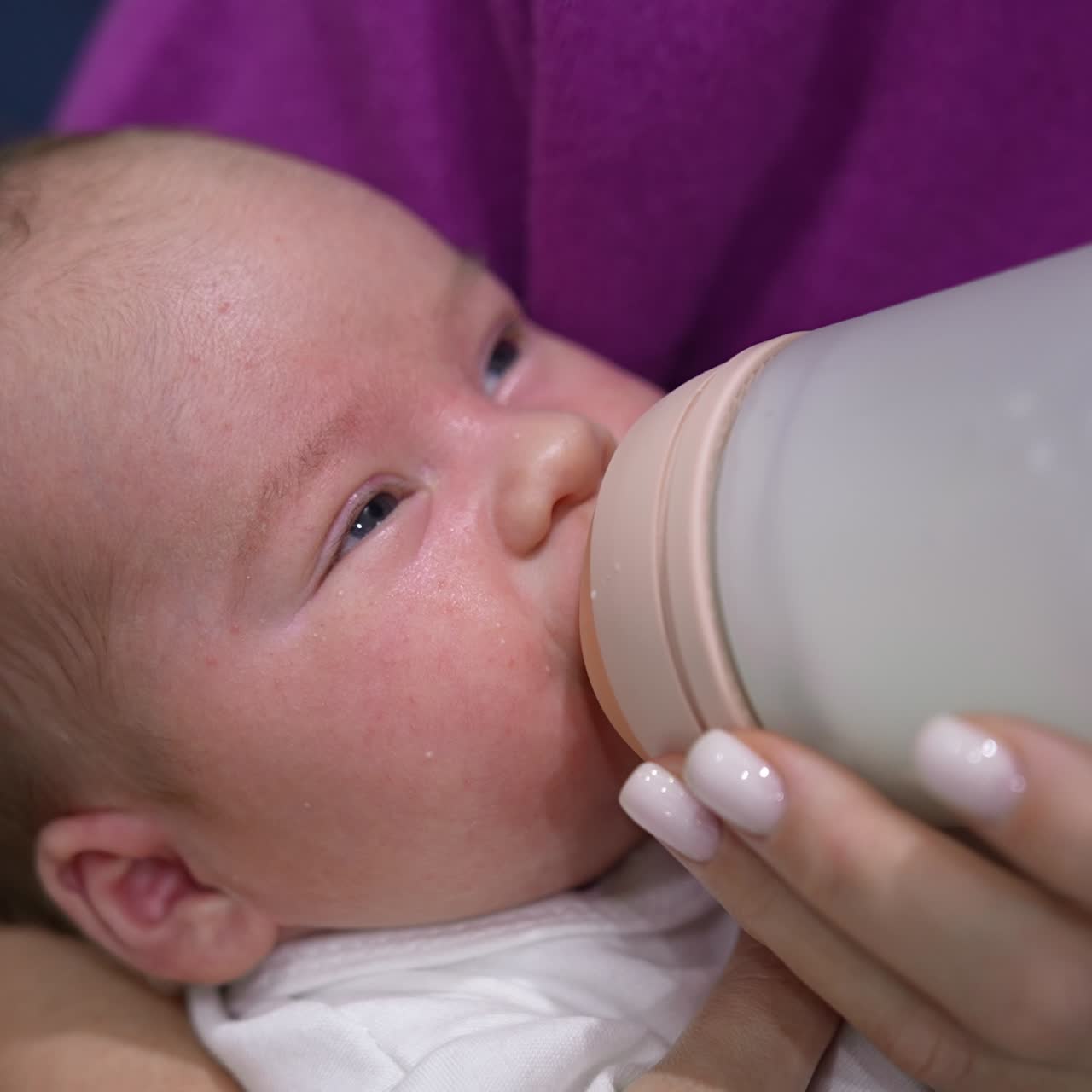 Newborn baby fed from a bottle lying in mother's hands. Baby boy screwing up his eyes against the light while being fed. Close up