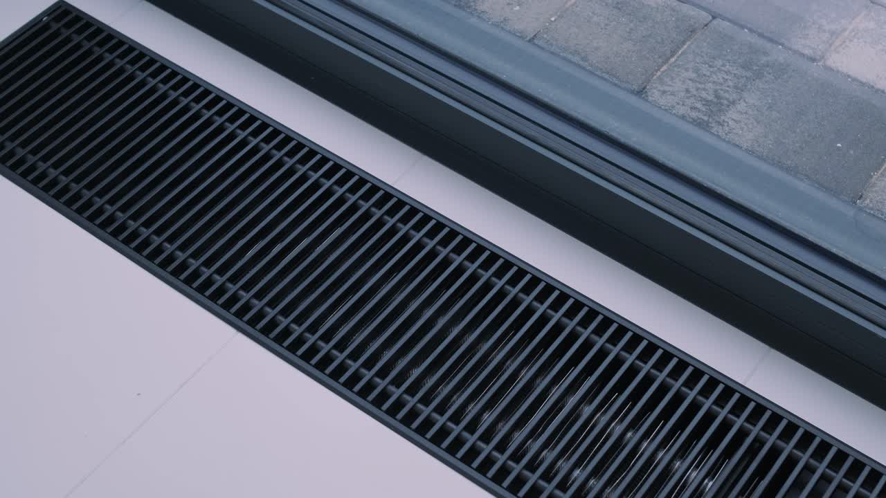 A modern linear floor vent providing efficient air circulation in a clean, white interior space, juxtaposed with a window view to the exterior gray bricks