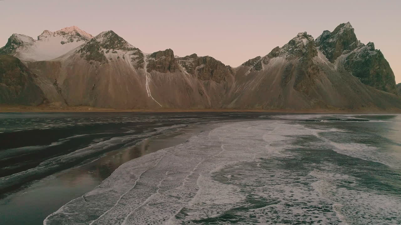 Rocky snow capped Vestrahorn Mountain and Stokksnes sunrise ocean waves aerial reversing view
