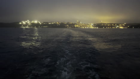 Viajar-En-Barco-Durante-La-Noche-Con-La-Tapa-De-La-Ciudad-En-El-Horizonte-Detrás-Del-Barco