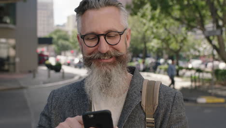 retrato de un hombre barbudo maduro y sofisticado sonriendo feliz usando un teléfono inteligente enviando mensajes de texto navegando en una concurrida calle de la ciudad