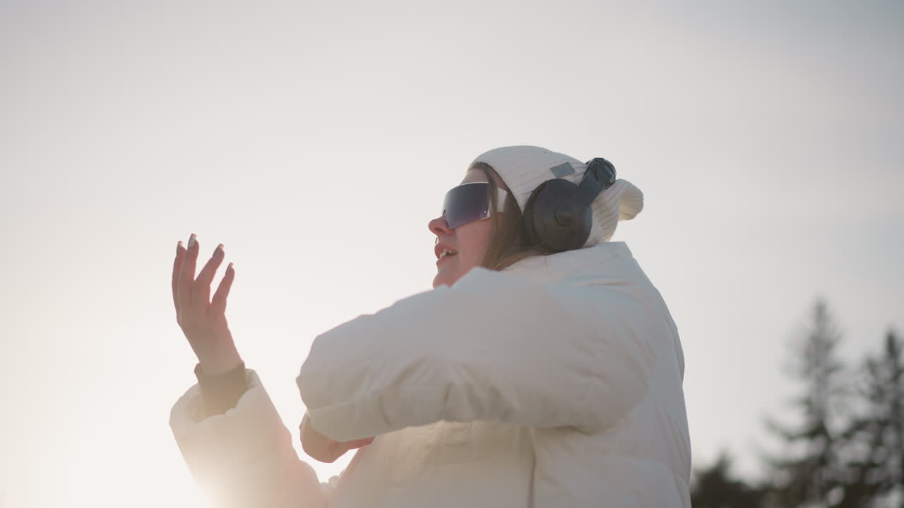 Young girl twirling freely with hands raised, smiling in winter coat, enjoying sunlight while wearing headphones and beanie, expressing joy and movement against soft sky and blurred trees