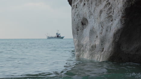 Sea-wave-gently-breaks-on-rocky-cliff-cave,-calm-Mediterranean-sea-with-cloudy-sky-and-boat