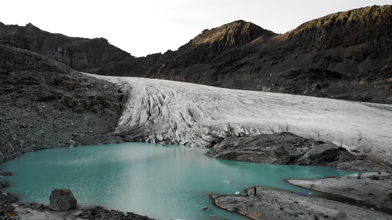 sobrevuelo aéreo sobre el agua turquesa del lago glacial en el glaciar hohlaub cerca de saas-fee en valais, suiza con un excursionista en la orilla disfrutando de la vista del hielo y los picos brillantes