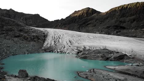 Aerialf-flyover-over-the-turquoise-water-of-the-glacial-lake-at-Hohlaub-glacier-near-Saas-Fee-in-Valais,-Switzerland-with-a-hiker-at-the-shore-enjoying-the-view-of-the-ice-and-glowing-peaks