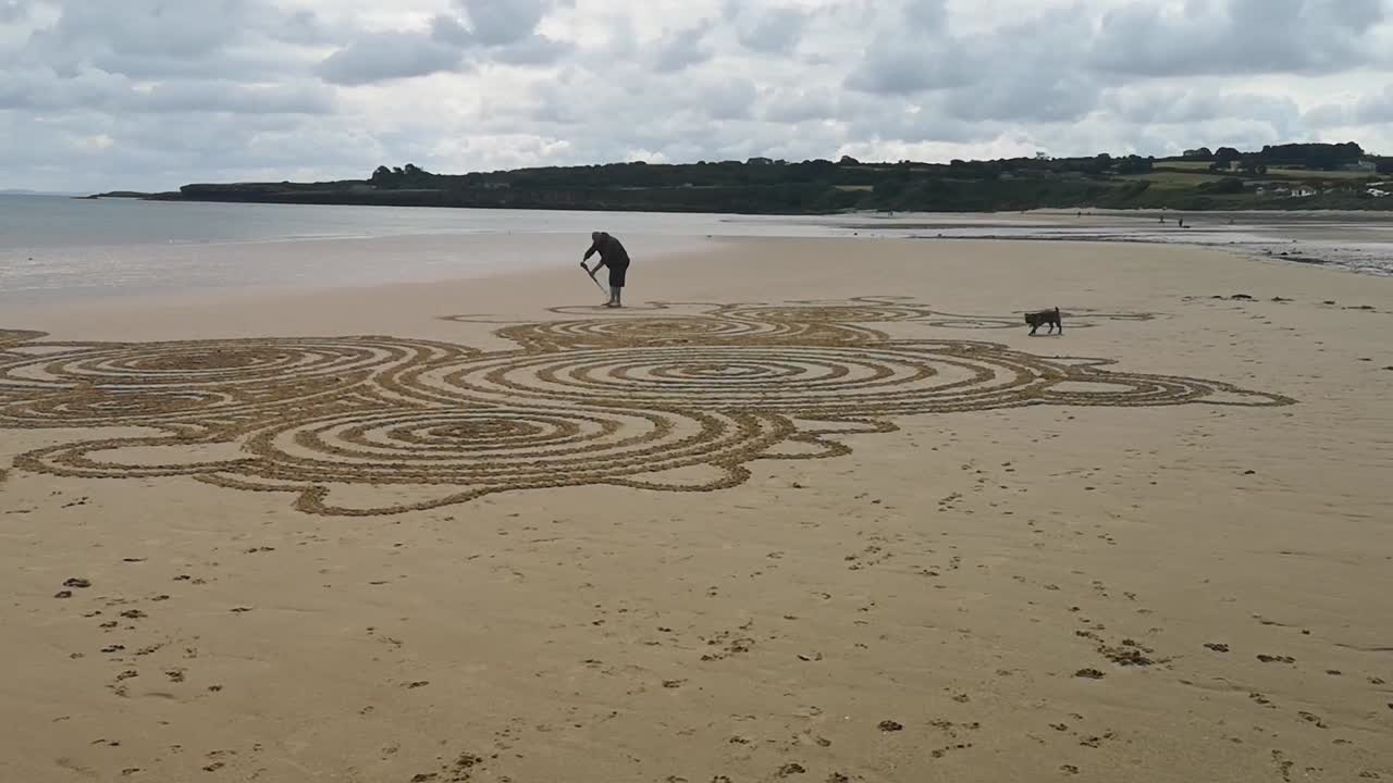 남성이 테리어 개와 함께 모래 앤글리 해변에서 예술적인 흐르는 패턴을 만들고 있습니다.
