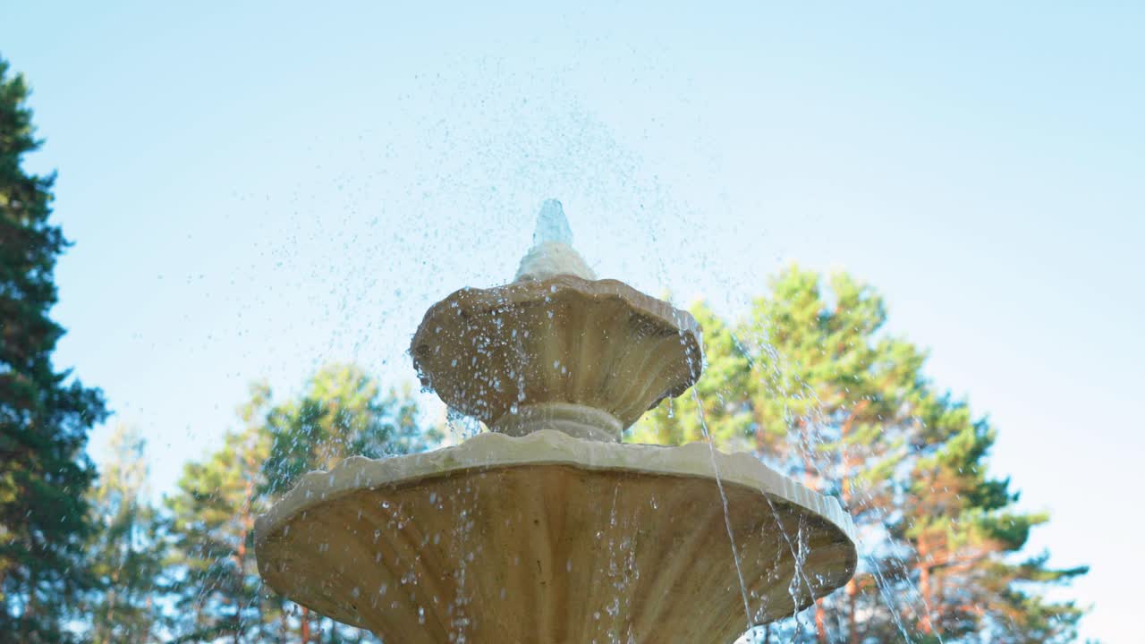 Low angle shot of a water fountain with dripping water drops in the park with green pine trees and blue sky in the background in summer day