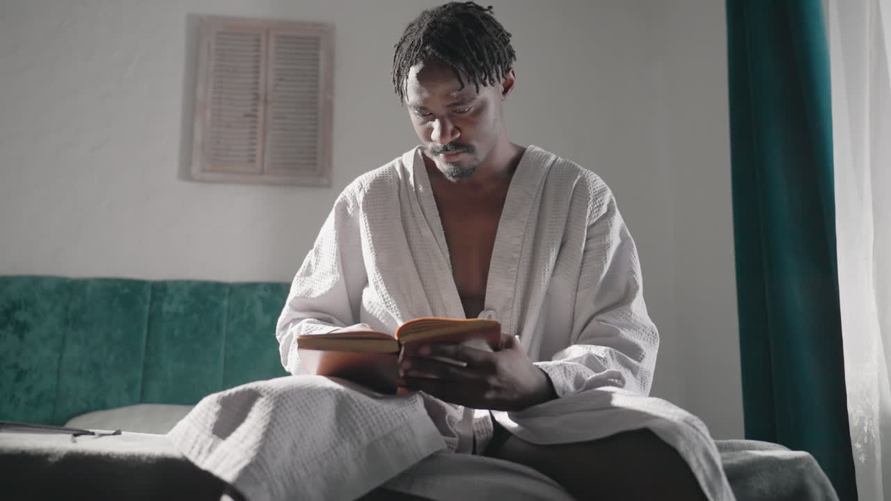 Man in bathrobe sitting on bed, reading book with focus. Bright morning light fills room, soft atmosphere. Quiet, cozy indoor scene with tranquil mood and relaxed vibes