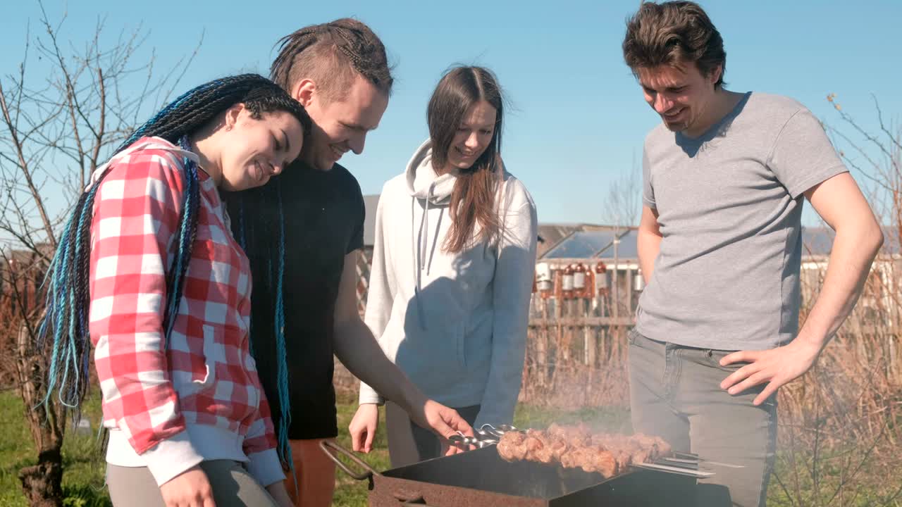 Group of young people friends Barbecue shashlik meat on top of charcoal grill on backyard. Talking and smiling together.