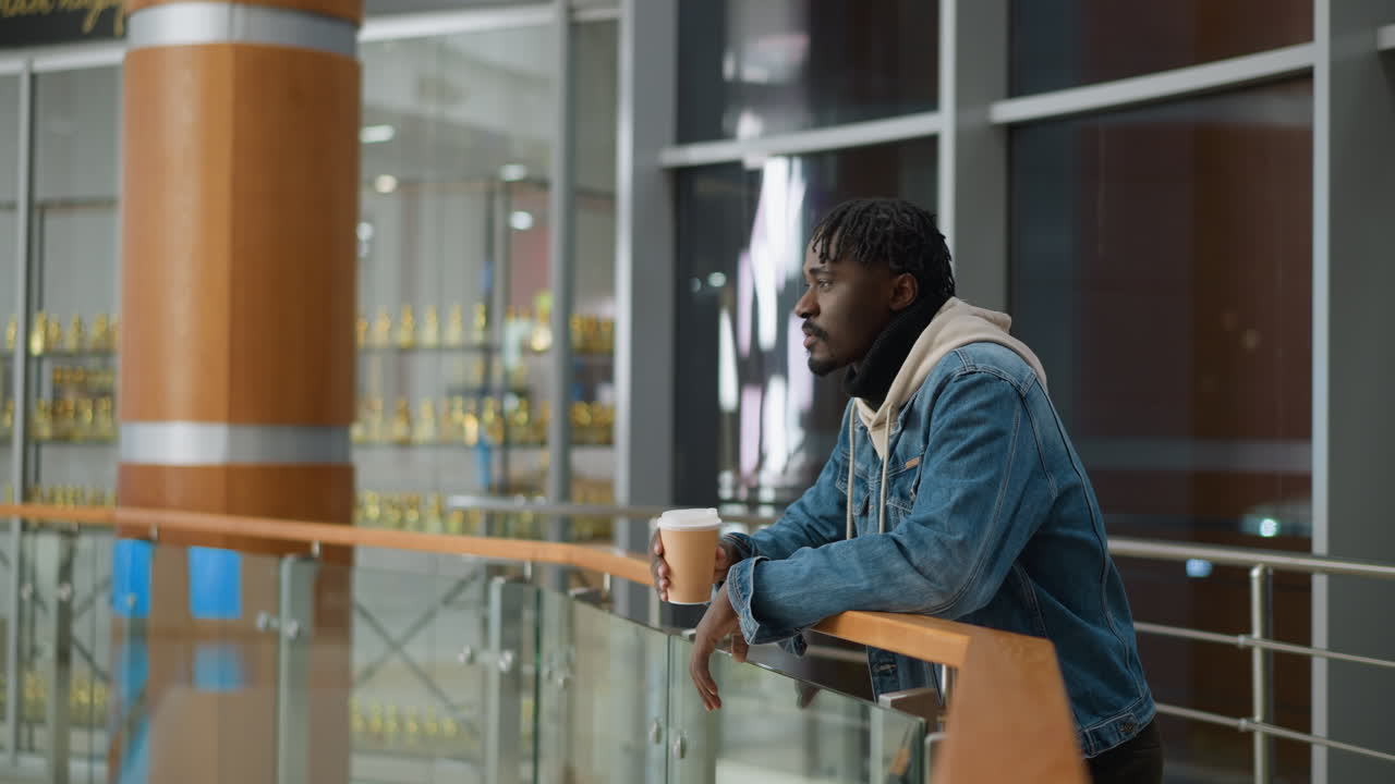 Young man in casual denim jacket and hoodie standing, holding paper cup, enjoying hot beverage while leaning on balcony railing in modern indoor space, contemplating and relaxing