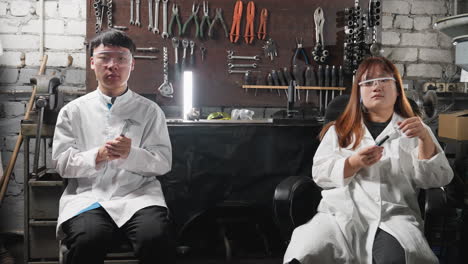 자동차 작업실에 앉아있는 중국 실험실 기술자, 안전 안경과 함께 베르니에 칼리퍼를 들고 있는 소년, 여성이 좌석을 부드럽게 회전시키는 동안, 도구와 장비가 벽에 매달린 산업 작업 공간