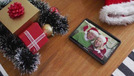 una pareja caucásica senior sonriente con sombreros de papá noel en una llamada de video de navidad en una tableta.