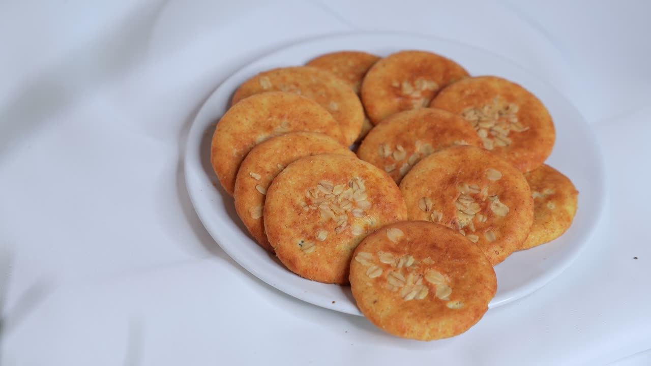 Close-up of a woman's hand picking a rustic oat cookie from a plate on a white tablecloth. Freshly baked snack, perfect for food, health, or lifestyle concepts. Filmed in crisp 4K.