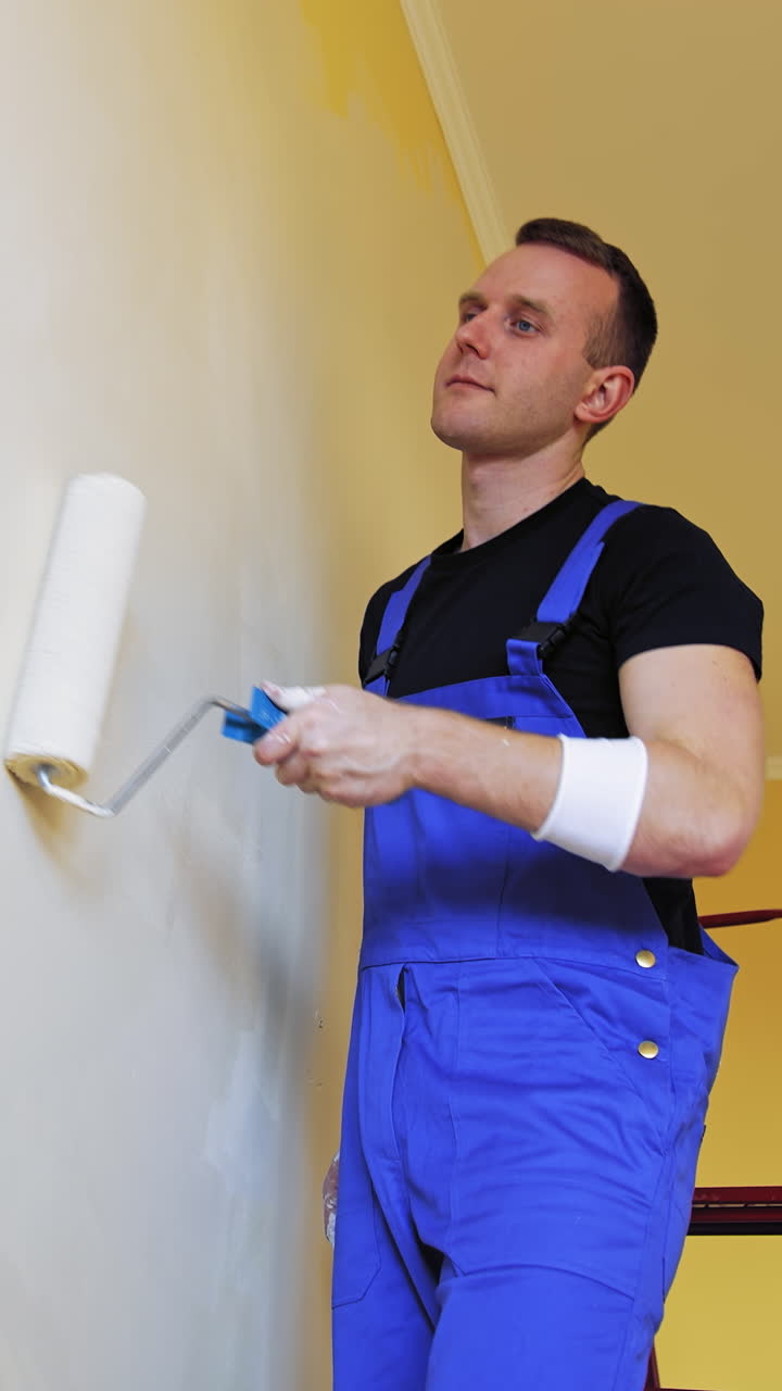 Painter man painting wall with a roller. Portrait of a young worker in overalls doing makeover with new color in the room. Professional painting services. Vertical video