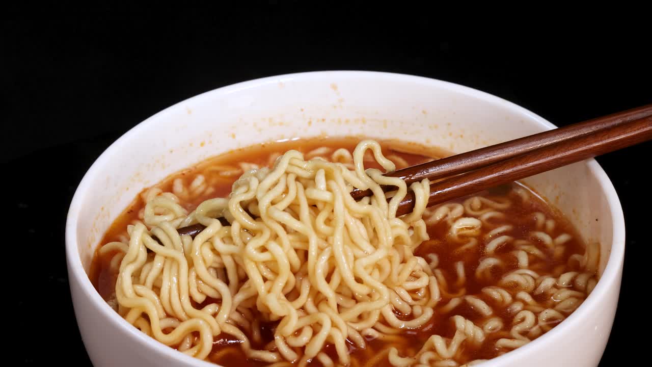Chopsticks grasp and lift cooked instant noodles from a steaming bowl of broth, captured in close-up with dramatic lighting and a dark background
