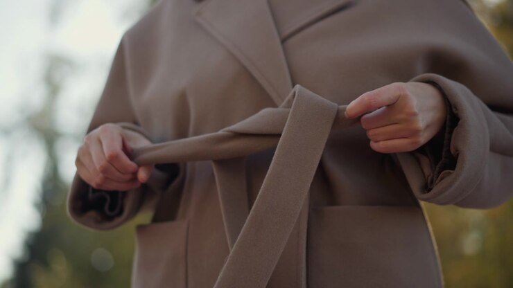Close up of woman tying belt on her brown coat focusing solely on hands and lower torso. action is deliberate as woman adjusts fabric texture details against outdoor backdrop with soft