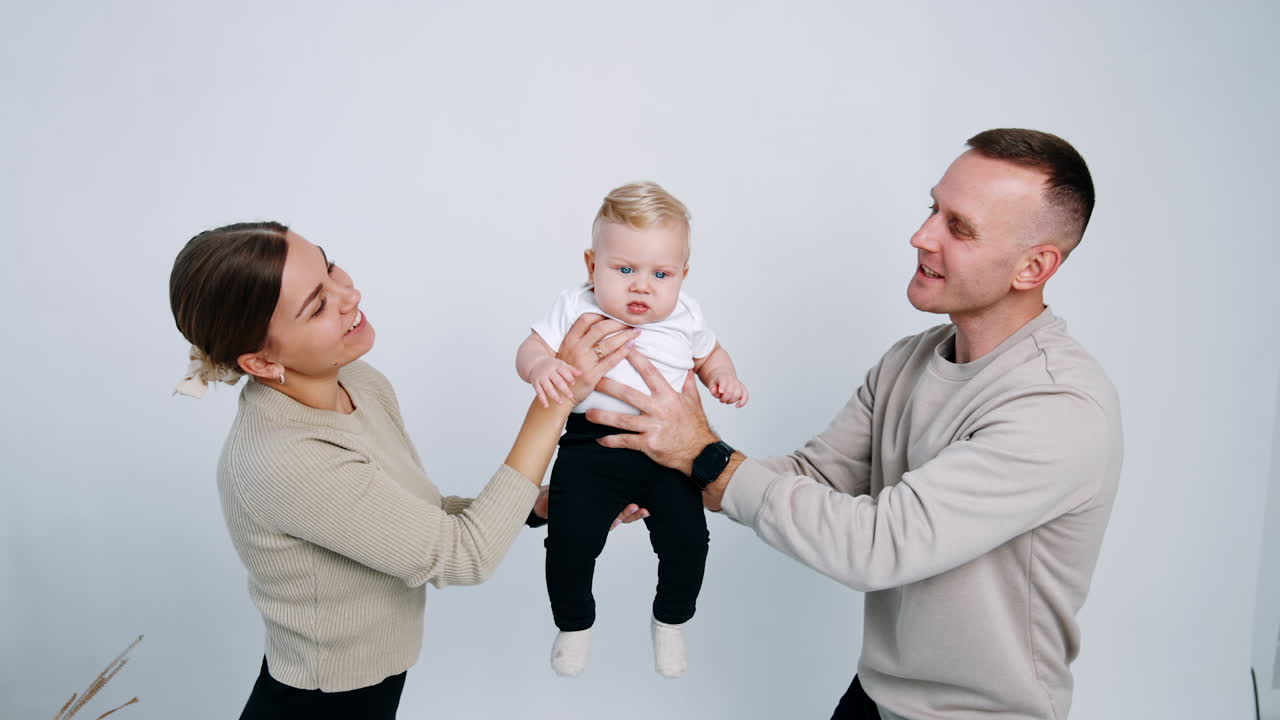 Mom and dad toss their baby holding him from two sides. Little kid waves his hands and starts to cry. White backdrop.