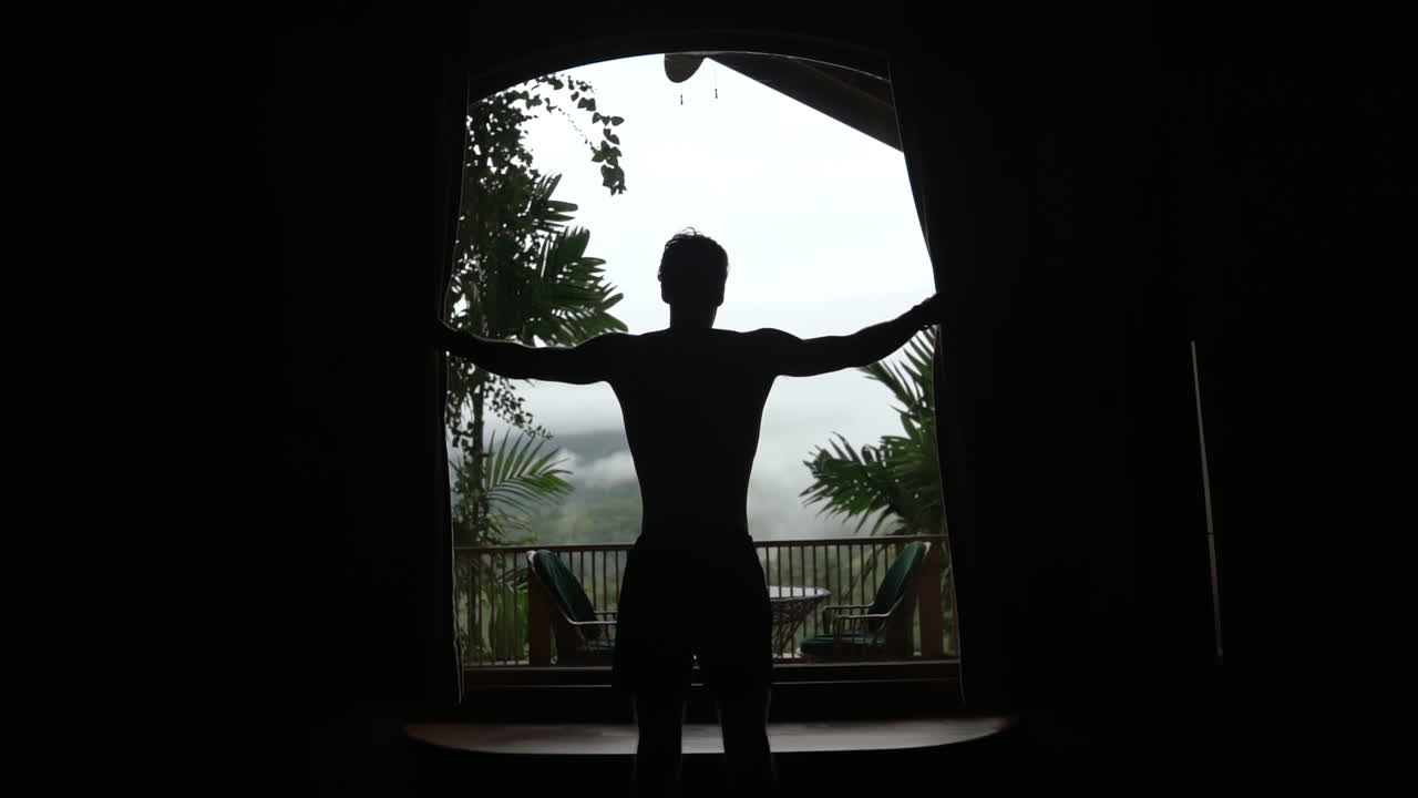 Silhouetted man opens blinds to reveal foggy rain forest balcony, slow motion