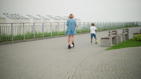 back view of mother and her son ride scooters side by side along a curved paved walkway, with iron railings on one side and benches along the path