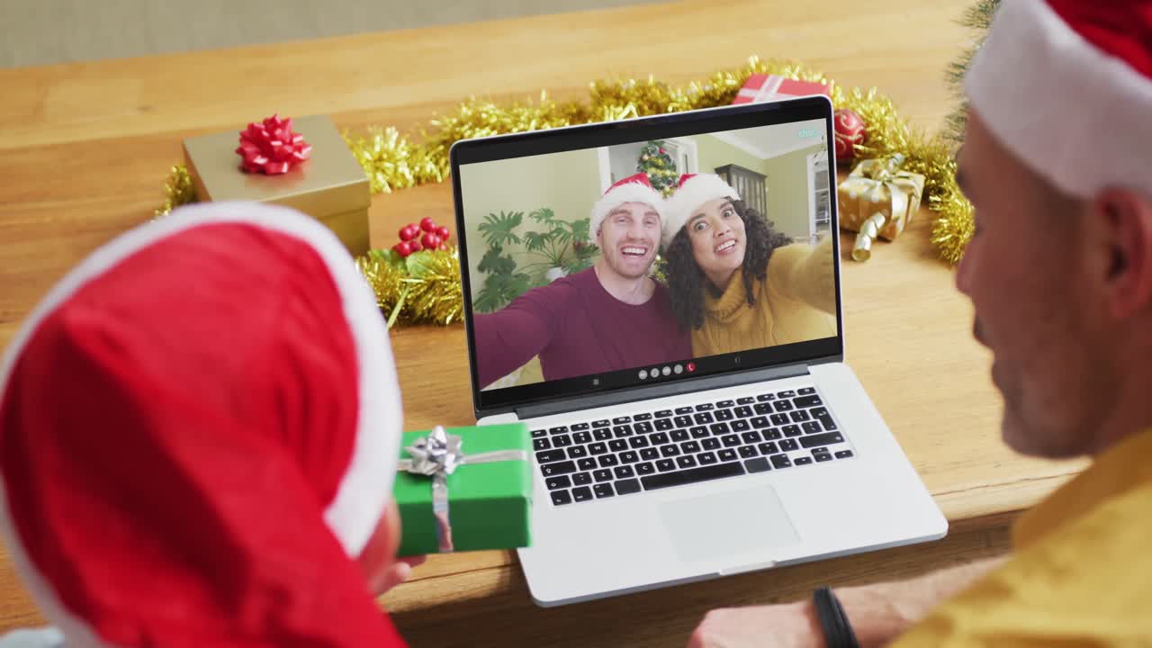 padre y hijo caucásicos con sombreros de santa usando una computadora portátil para una videollamada de navidad con una pareja en la pantalla