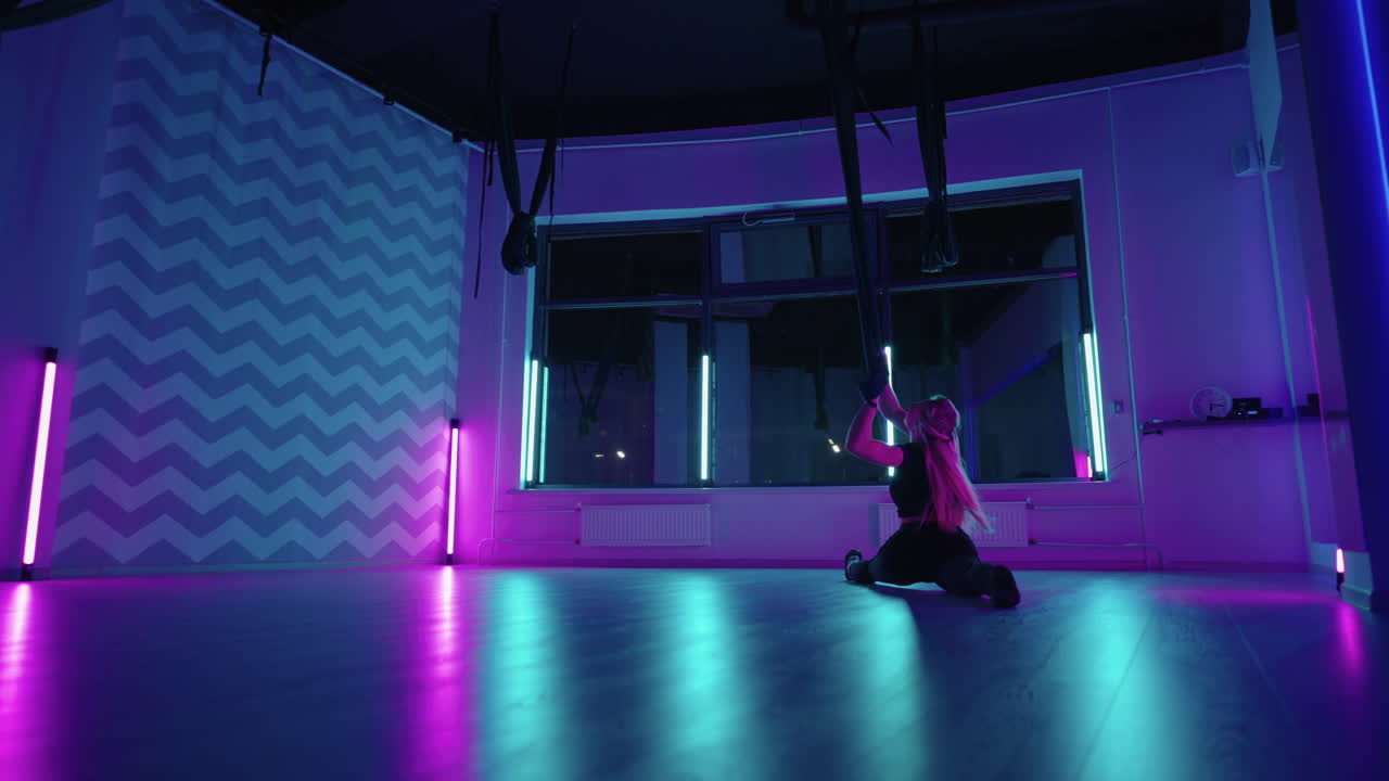 A woman does yoga and stretching on a hanging hammock in neon light. Young pretty slim body fitness girl practicing fly yoga in the gym.
