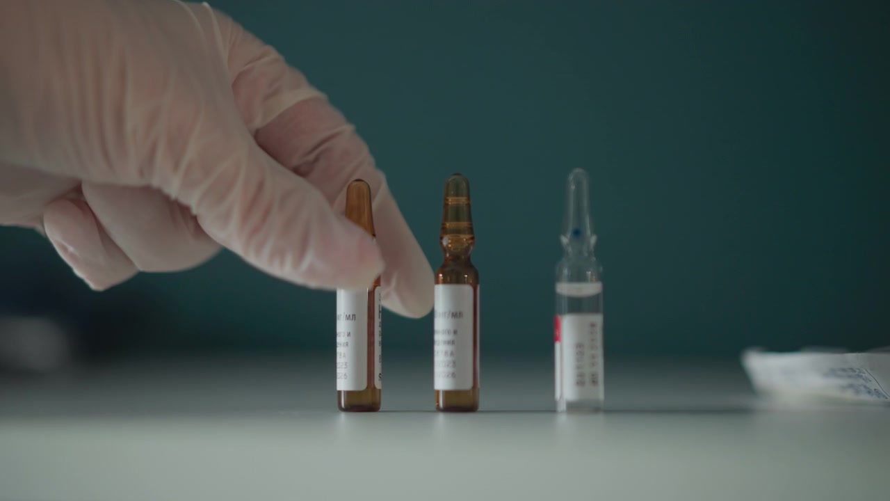 Gloved hand selects amber ampoules, tense clinic lighting and labeled vials on white surface, sterile gauze nearby, careful dosing gesture by medical technician preparing medication