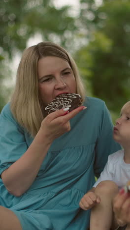 una madre disfruta de un bocado de chocolate mientras está sentada en el suelo bajo un árbol con sus dos hijos, un niño la mira con curiosidad mientras come, mientras el otro niño juega