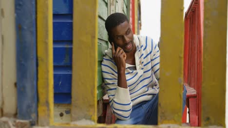 front view of young black man talking on mobile phone sitting on stairs of hut at beach 4k