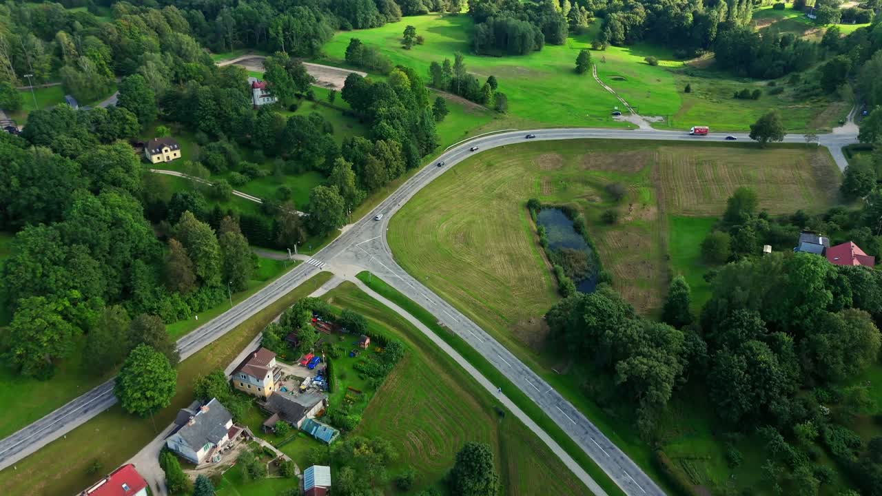 Cars are driving through an intersection in Talsi, Latvia, surrounded by houses, trees, and green fields, creating a picturesque countryside scene